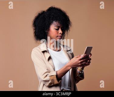 A young woman with curly hair, wearing a beige jacket and white shirt, looks intently at her smartphone, standing against a plain neutral background. Stock Photo