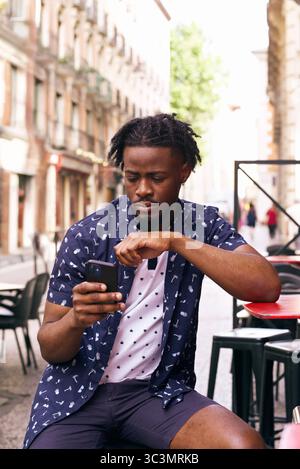 A black man sits at a bar terrace, deeply engaged with his smartphone. The street is lively, adding a vibrant city atmosphere to this casual urban sce Stock Photo