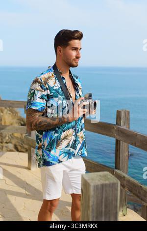 A young man stands confidently under the bright sun, holding a camera by the seaside. He exudes a sense of adventure and relaxation, dressed casually Stock Photo