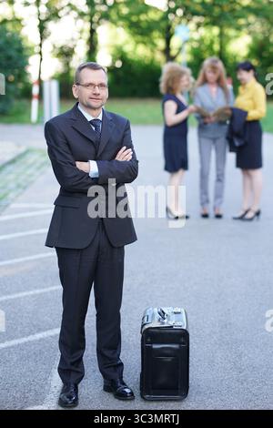 male boss in suit stands at a distance from his female secretaries, arms crossed Stock Photo