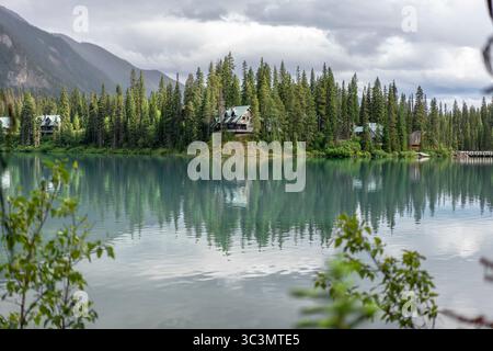 A serene lakeside view in Canada with a cozy house nestled among tall evergreen trees. The calm water reflects the natural beauty, creating a peaceful Stock Photo