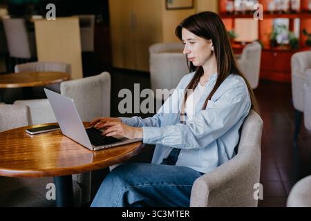 A woman is comfortably working on her laptop in a cozy cafe. She is seated at a wooden table, focused on her tasks, surrounded by a warm, inviting amb Stock Photo
