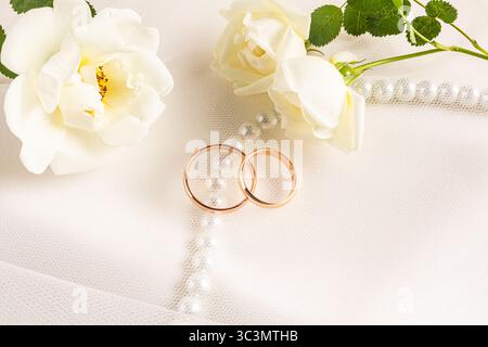 Elegant gold wedding rings rest on luxurious beige satin, surrounded by delicate white roses and pearls, creating a romantic and timeless scene of bea Stock Photo