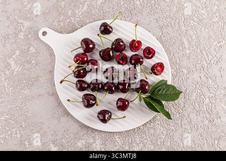 Top view of juicy ripe cherries elegantly arranged on a white cutting board plate. The textured gray stone background enhances the colorful and fresh Stock Photo