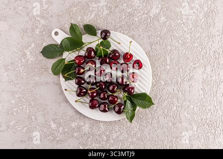 Top view of juicy ripe cherries scattered on a white  cutting board plate adorned with green leaves, set against a textured light stone background, cr Stock Photo
