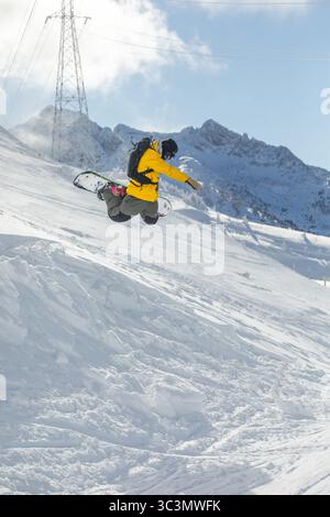 A man in a yellow jacket performs an exhilarating freeride snowboarding maneuver on a snow covered mountain, under a clear blue sky, capturing the thr Stock Photo