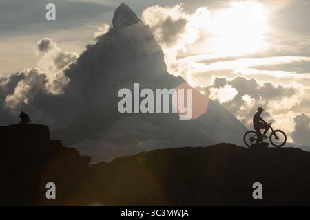 Unrecognizable silhouette of a man standing on a bike on a rocky alpine trail, set against the dramatic backdrop of sunlit clouds and the famous Matte Stock Photo
