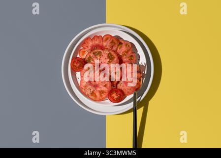 Top view of a vibrant raf tomato cut in half, sprinkled with salt and spices on a white plate with fork against a yellow and gray background, highligh Stock Photo