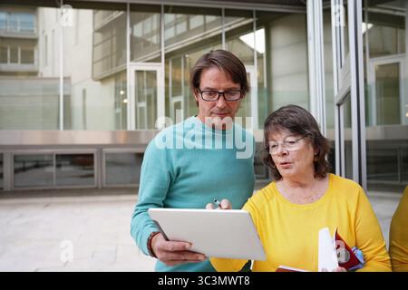 An elderly teacher and a student engage in a learning session outdoors, using a digital tablet and printed notebook, showcasing collaboration and mode Stock Photo