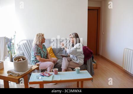 Two senior female friends sit comfortably at home, engaging in a warm and meaningful conversation. The cozy setting emphasizes their close bond and re Stock Photo