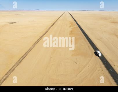 Aerial view of a lone vehicle travels on a straight road cutting through the vast desert landscape under a clear blue sky, Walvis Bay, Walvis Bay, Namibia. Stock Photo
