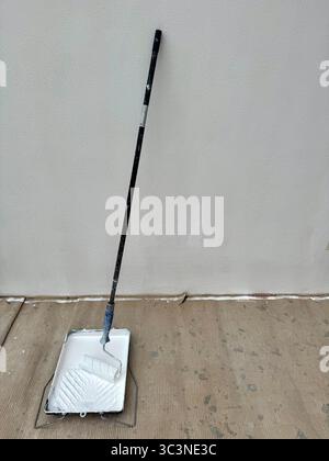 Paint roller with an extension pole resting in a tray filled with white paint, placed on a protective floor covering in front of a wall being painted. Stock Photo
