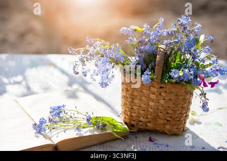 Bright blue forget-me-not flowers in a basket next to an open book on a sunny day at a garden table. Stock Photo