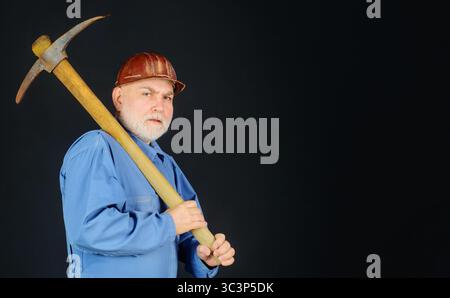 Serious builder in uniform with pickaxe on shoulder. Coal miner with pickax. Male laborer with hand tool for digging, mining. Construction worker in Stock Photo