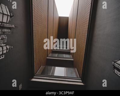 Narrow view looking up between two high brick walls in orange tones, flanked by dark walls. Several windows with reflective glass are embedded along t Stock Photo