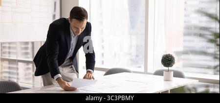Businessman leaning over table, carefully examining papers with data Stock Photo