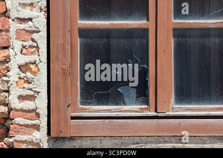 A close-up of a cracked glass window in a wooden frame set into a rough brick wall reveals damage and the aging condition of the structure’s exterior Stock Photo