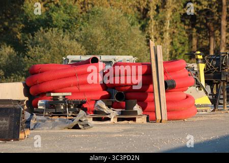 Bright red flexible utility pipes are coiled on an asphalt surface at a construction site, intended for upcoming underground conduit installations Stock Photo