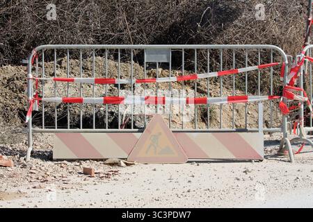 A metal barrier with red and white warning tape and a faded work sign blocks road access, warning of ongoing construction and ensuring site safety Stock Photo