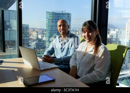 Two multiracial business colleagues sitting in the office desk, looking at camera smiling. Stock Photo