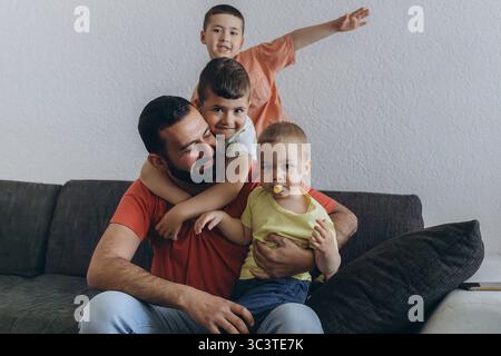 Happy middle eastern father having fun with his three sons at home. Dad cuddling toddler while other kids play and smile. Loving family moment on cozy Stock Photo
