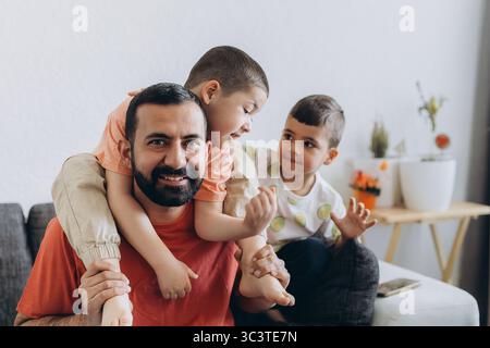 Happy middle eastern father having fun with his three sons at home. Dad cuddling toddler while other kids play and smile. Loving family moment on cozy Stock Photo