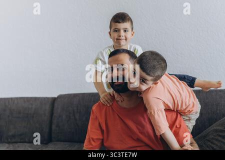 Happy middle eastern father having fun with his three sons at home. Dad cuddling toddler while other kids play and smile. Loving family moment on cozy Stock Photo