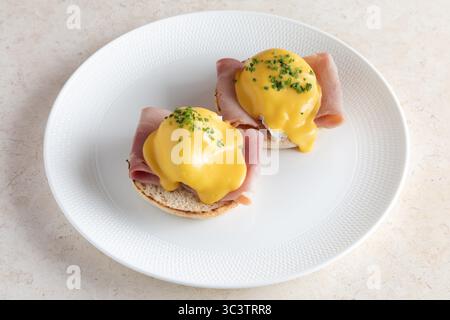Classic eggs Benedict with poached eggs, creamy hollandaise sauce and ham on English muffins. Gourmet breakfast dish with fresh chive garnish. Stock Photo