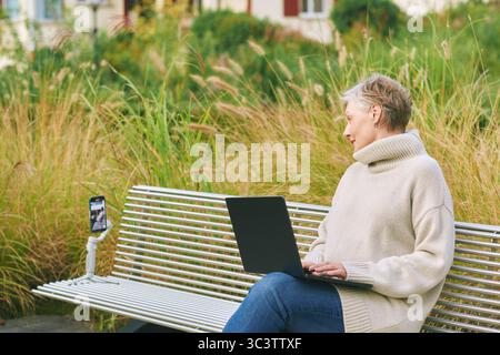 Outdoor portrait of happy and active mature woman recording herself with phone while using laptop, social media and vlogging concept Stock Photo