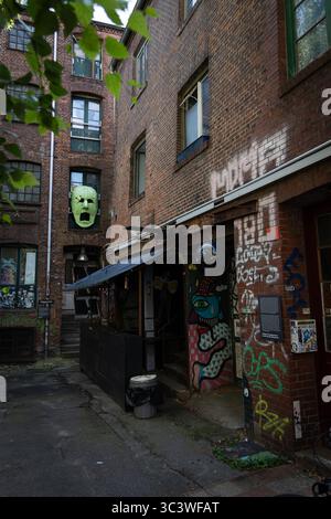 Urban scene of an old window covered with various stickers, set in a graffiti-covered brick wall, with pink flowers and green plants growing below. Stock Photo