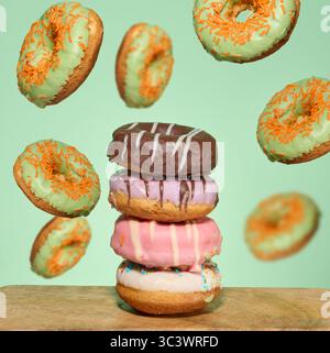 A stack of four brightly glazed donuts on a table with green donuts with orange sprinkles flying around them Stock Photo