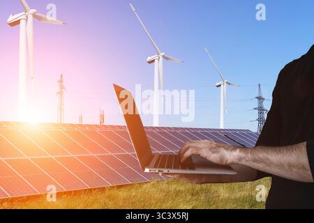 Modern sustainable energy concept. A technician or engineer uses a laptop against the backdrop of solar panels and wind turbines during a sunny day. The image illustrates clean energy monitoring, smart grid technology, and renewable power infrastructure. Perfect for topics such as green technology, energy transition, digital transformation in utilities, and environmental innovation. Stock Photo