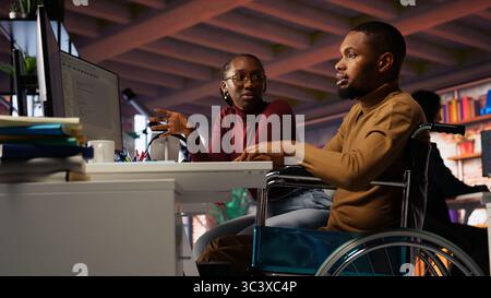 Manager coming to inclusive startup office employee in wheelchair desk to offer help. Thoughtful supervisor checking up on software development agency worker with disability, camera B Stock Photo