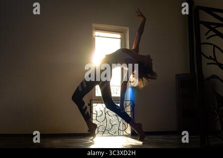 Slim sporty woman doing back band dancer pose in front of window, where come golden sunlight. Morning yoga routine, dancing, self care, active Stock Photo