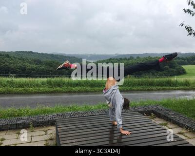 Unrecognisable young woman doing hand stand with wide open legs like split in front of green nature environment. Active life style, yoga, gymnastic. Stock Photo
