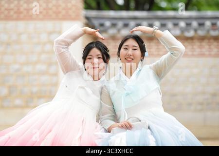 In June in Seoul, two Korean women in their twenties sit on a grassy plaza at Gyeongbokgung Palace, making a heart with hands while wearing cute pink Stock Photo