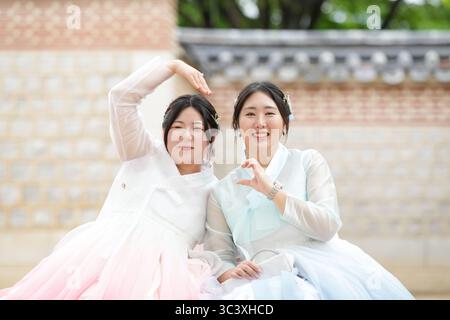 In June in Seoul, two Korean women in their twenties sit on a grassy plaza at Gyeongbokgung Palace, making a heart with hands while wearing cute pink Stock Photo