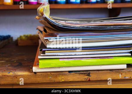 A colorful stack of sketchbooks, drawing pads, paper portfolios,  layers of paper, canvas boards, paint tubes, and mixed media folders on a wooden des Stock Photo