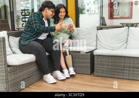 Happy latin couple using smartphones while sitting on an outdoor sofa, woman with prosthetic leg Stock Photo