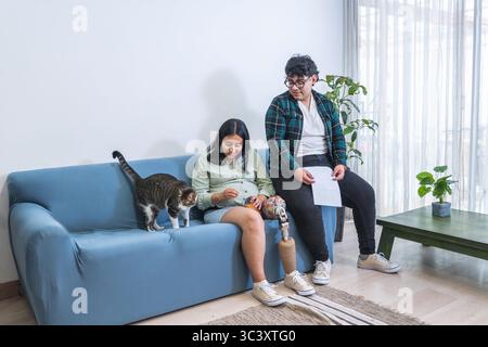 Pregnant woman with prosthetic leg sitting on sofa with partner playing and cat Stock Photo