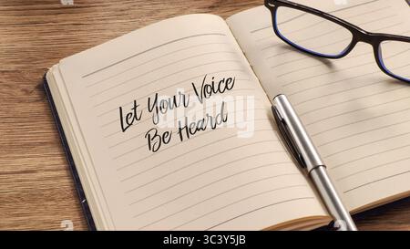 Opened notebook on a wooden desk with a pen and glasses, featuring the handwritten motivational phrase "Let Your Voice Be Heard" on the page. Stock Photo