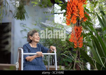 Smiling elderly woman in garden. Happy senior woman sitting on terrace with red jade vine flowers at home. Stock Photo