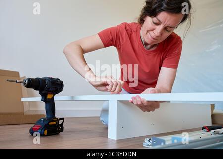 Woman assembling bed frame on wooden floor, using wrench and drill for installation. DIY furniture assembly and home improvement Stock Photo