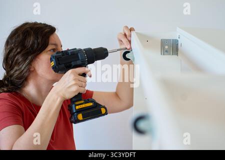 Woman using cordless drill to screw part into white bed frame. Female assembling furniture in new apartment. DIY home renovation and home organization Stock Photo