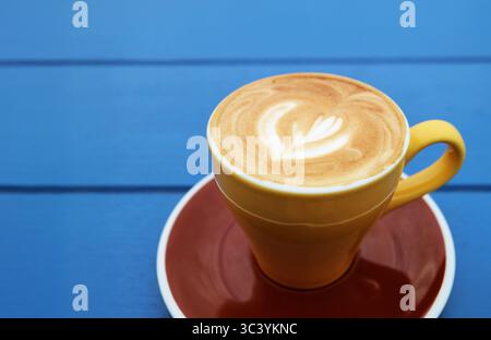 Frothy cappuccino coffee in yellow brown cup isolated on blue wooden table Stock Photo