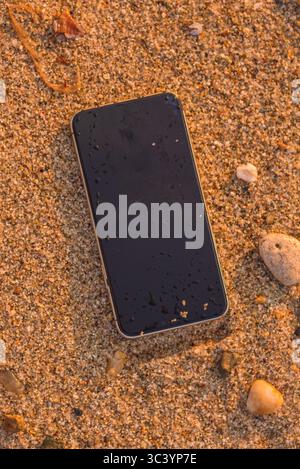 Wet smartphone lying on sand at beach with seashells and stones, symbol of carelessness, loss, or vacation mishap. Top view. Stock Photo