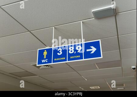A blue sign with white arrows hanging on the ceiling in a public area indicating toilets, stands and seating areas. Stock Photo