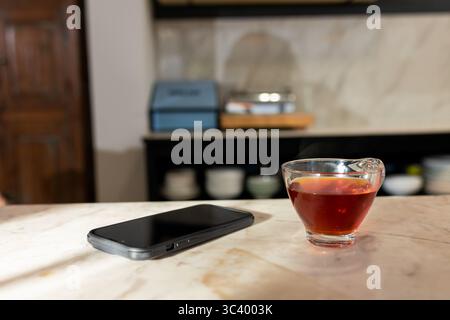 Smartphone lying beside cup of dark tea on marble countertop, with blurry bowls and boxes behind Stock Photo