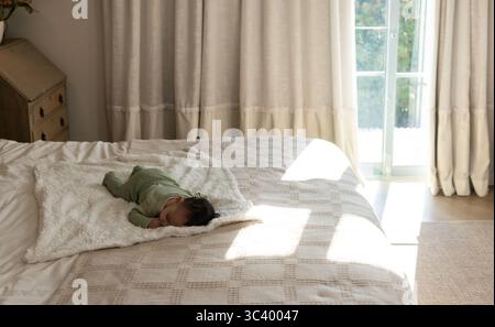 Infant girl lying on plush white throw on patterned coverlet in bedroom wearing pale green onesie Stock Photo