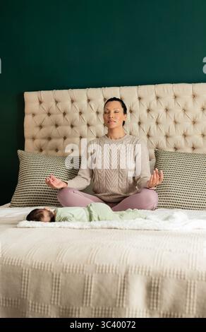 Mother and baby sitting on bed in bedroom meditating with tufted headboard and soft blanket Stock Photo